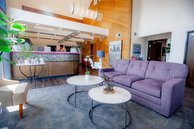 Bright modern lobby with purple sofa, marble coffee tables, wooden panel wall, and receptionist desk area.