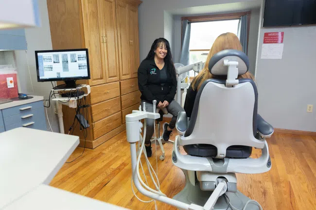 Dental hygienist smiling and talking to patient seated in dental chair in a bright clinic room.