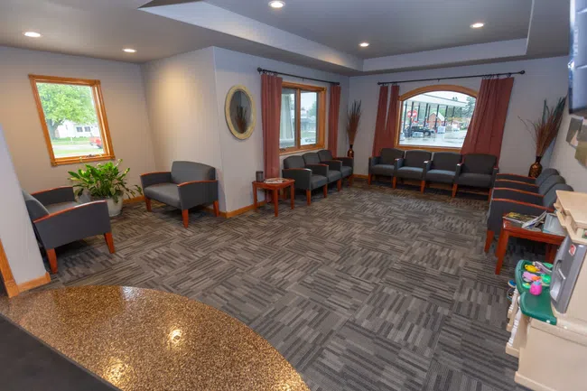 Waiting room with gray chairs, patterned carpet, large windows, and reddish curtains in a well-lit space.