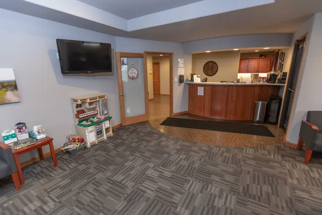 Reception area with wooden desk, wall-mounted TV, children's play area, and patterned carpet flooring.