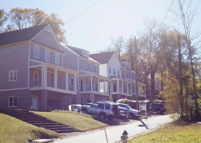Newly constructed two-story townhouses with front porches on a sunny residential street with parked vehicles.