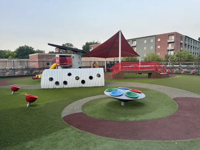 Modern outdoor playground with colorful equipment, shade canopy, and soft artificial turf surrounded by buildings.