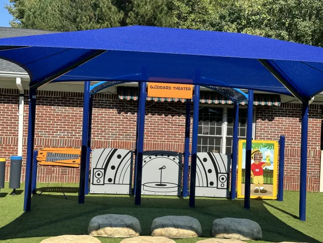 Outdoor children's play stage with blue canopy, labeled Goddard Theater, featuring a microphone and seating stones.