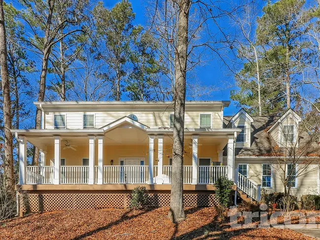 Large two-story beige house with a covered front porch surrounded by tall trees under a clear blue sky