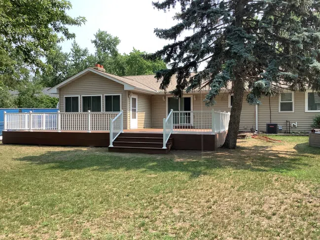 Single-story beige house with large wooden deck and white railings in spacious backyard with green lawn and trees