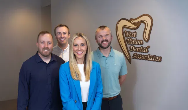 Four dental professionals smiling inside West Union Dental Associates office with logo on wall.
