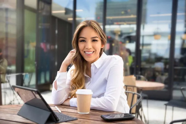 Smiling woman with long hair in white shirt sitting at outdoor cafe table with tablet, phone, and coffee cup