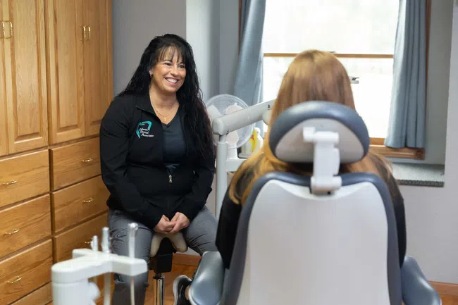 Female dental professional smiling and talking to a patient seated in a dental chair in a bright office.
