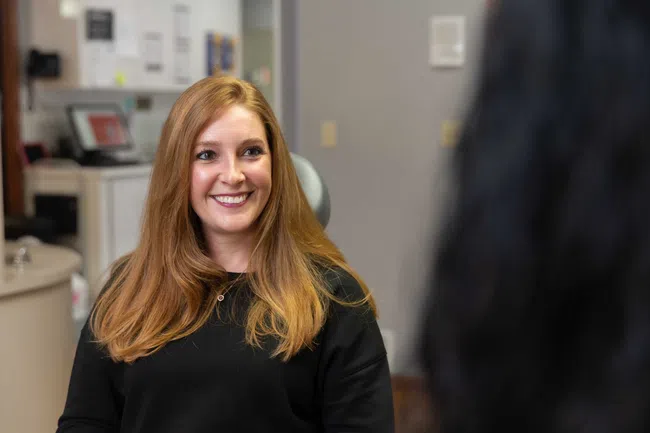 Smiling woman with long red hair wearing a black top sitting indoors facing another person.
