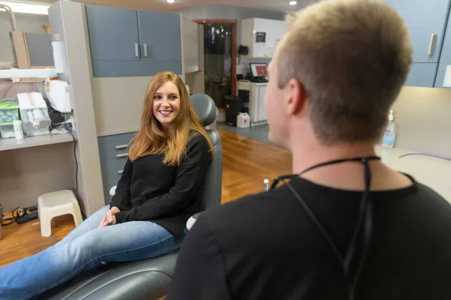 Smiling woman sitting in dental chair facing a dentist in a modern clinic with wooden floors.