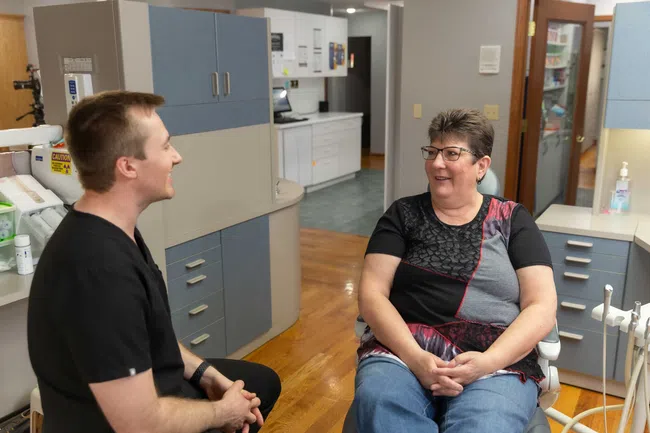 Dentist speaking with female patient seated in dental office wearing casual clothes and glasses