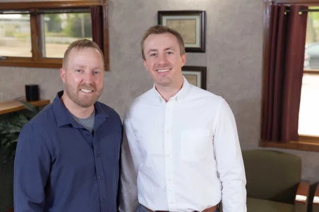 Two smiling men in business casual shirts posing indoors with framed pictures and windows in the background
