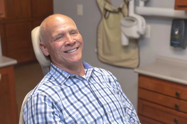 Smiling elderly man in checkered shirt sitting in dental chair in a modern dental clinic.