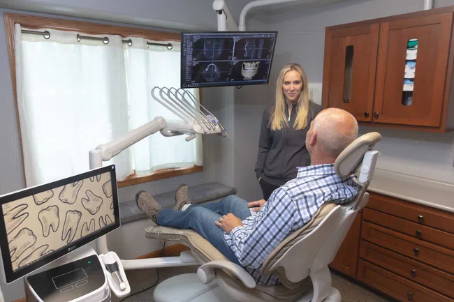 Dentist consulting a male patient in a dental office with X-ray images on two monitors in the background