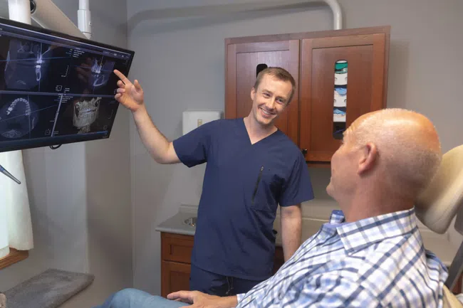 Dentist in blue scrubs showing dental X-rays on screen to an older male patient in a dental office.