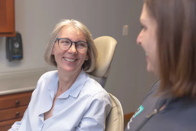 Smiling senior woman wearing glasses sitting in a dental chair talking to a female dentist.