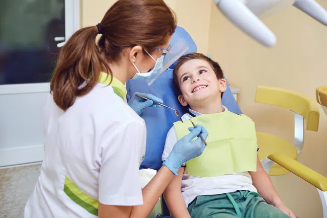 Child sitting in dental chair smiling at female dentist wearing gloves and mask during checkup.