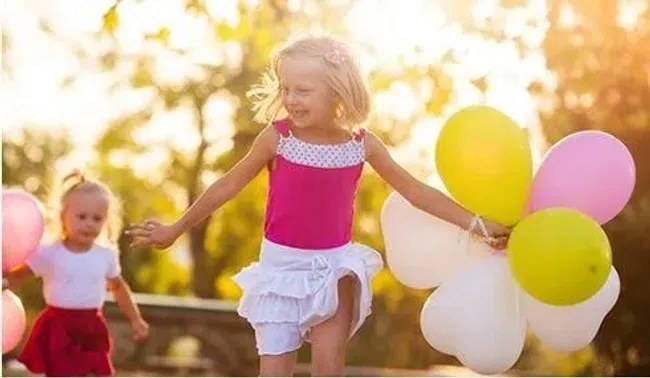 Two young girls playing outdoors holding colorful balloons on a sunny day with trees in the background