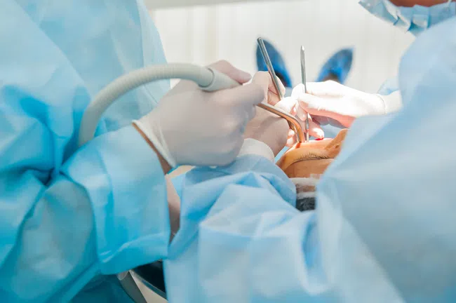 Dentists wearing blue surgical gowns perform dental procedure using dental instruments on patient.