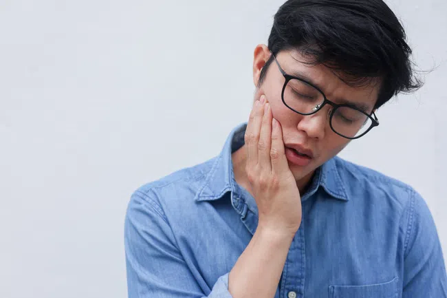 Young man in glasses and blue shirt touching his cheek with a painful expression against a white background
