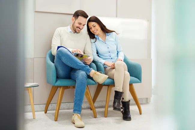 Smiling couple sitting closely on blue chairs reading a magazine in a bright modern waiting room.