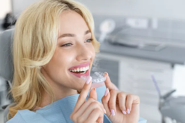 Smiling woman holding a clear dental aligner tray in a modern dental clinic setting.