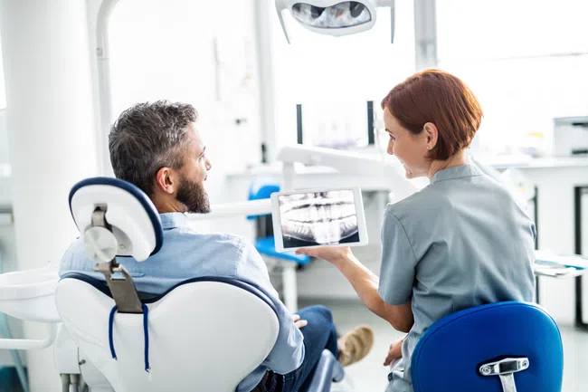 Dentist showing a male patient a dental X-ray image on a digital tablet in a bright dental office.