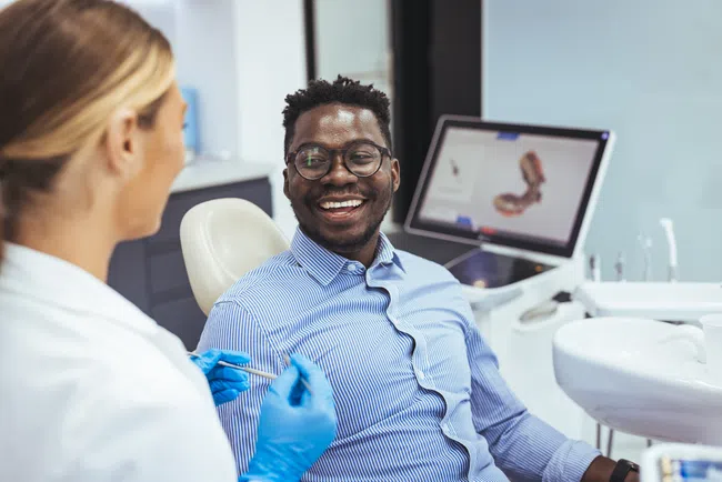 Smiling man sits in dental chair talking with female dentist wearing blue gloves in modern clinic.