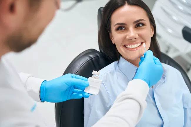 Dentist matching dental veneers shade to smiling woman at dental clinic during consultation.