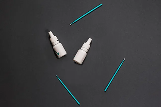 Two white nasal spray bottles and four blue cotton swabs arranged on a dark gray background.