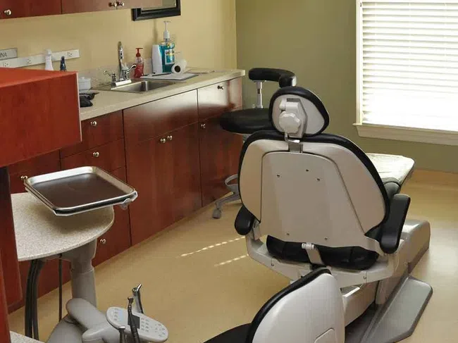 Modern dental office with patient chair, overhead light, cabinets, sink, and a TV mounted on the wall.