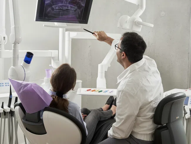 Dentist explaining dental X-ray results to a patient sitting in a dental chair with equipment around.