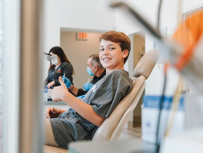 Smiling boy with braces gives thumbs up while sitting in dental chair with masked staff in background