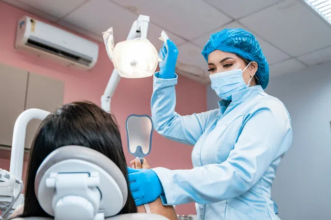 Dentist wearing blue scrubs and mask adjusting dental light while patient holds mirror in dental clinic.