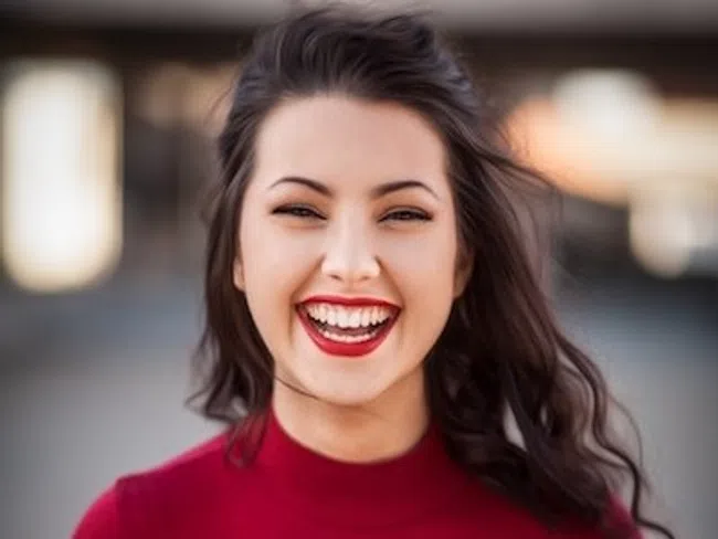 Smiling young woman with dark hair in a red top standing outdoors with blurred background.