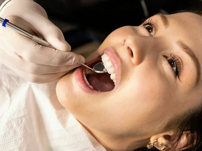 Dentist in gloves examining young woman's teeth with dental mirror during orthodontic checkup.