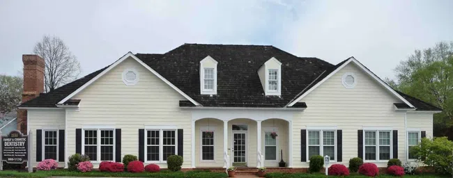 Large white family dentistry office building with black shutters, green shrubs, and pink flowers in front.