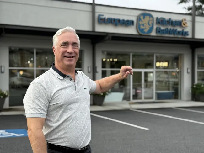 Smiling man in gray polo shirt pointing at European Kitchen & BathWorks storefront with glass windows