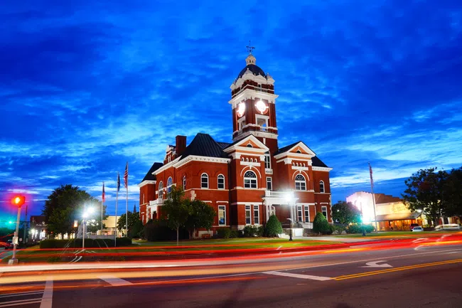 Historic red brick courthouse with clock tower illuminated at dusk and light trails from passing traffic.