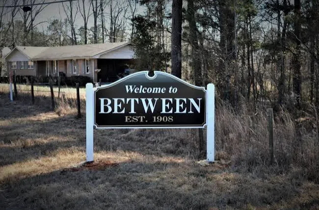 Welcome to Between town sign in front of rural house and trees, with overlaid text reading Welcome to BETWEEN EST. 1908