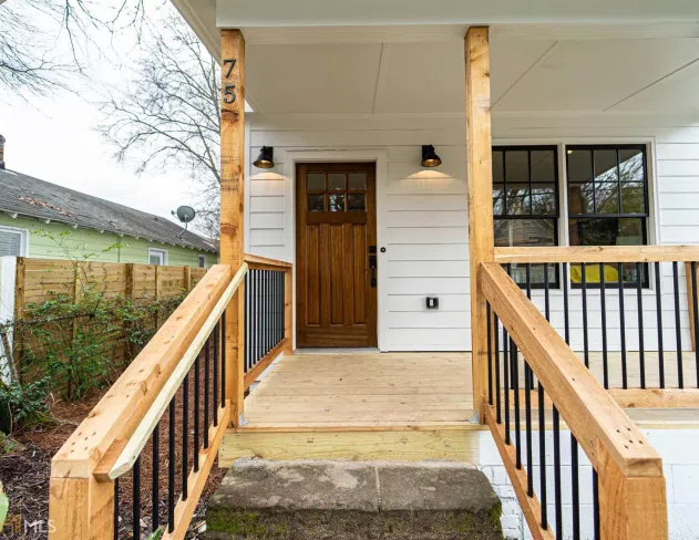 Front porch of a house with wooden railings, a wooden door, white siding, and outdoor wall lights.