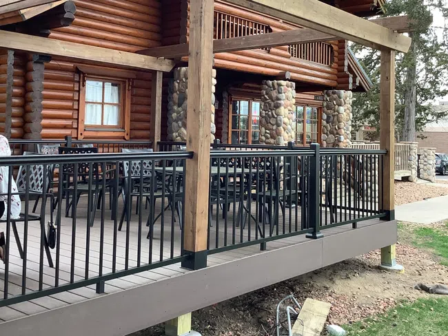 Log cabin porch with wooden beams, stone pillars, black metal railing, and outdoor seating under trees.