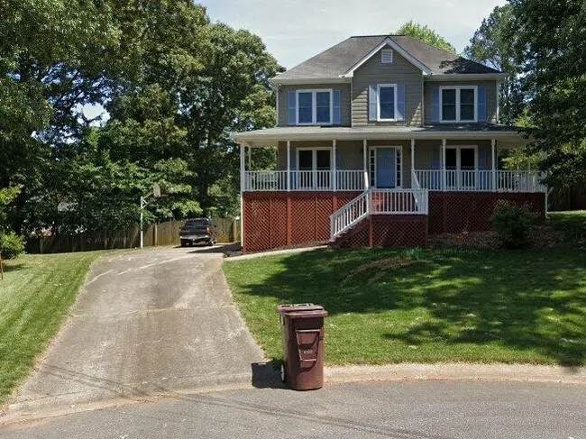 Two-story gray house with large front porch, driveway, green lawn, and trees on a sunny day.
