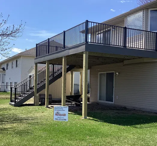 Elevated wooden deck with black metal railing attached to a beige two-story house under clear blue sky.