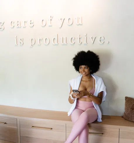 Young woman in pink activewear sitting on a wooden bench, holding phone, with wellness quote on wall behind her.