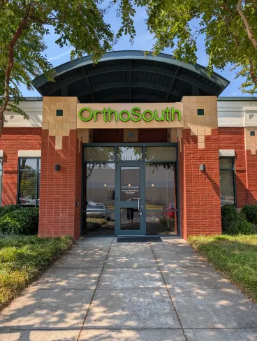 Entrance of OrthoSouth clinic with green signage, red brick walls, glass door, and surrounding greenery under blue sky.