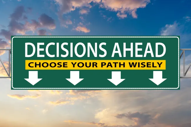 Green road sign with text 'Decisions Ahead, Choose Your Path Wisely' against a colorful sky background.