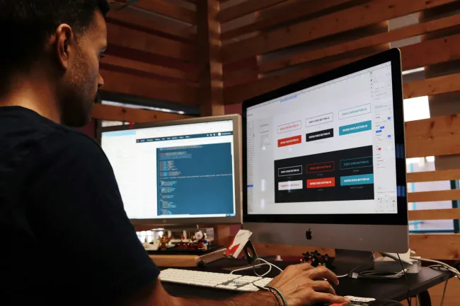 Man coding and designing buttons on dual monitors in a wooden office setting.