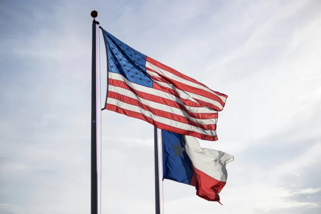 American and Texas flags waving against a cloudy sky, symbolizing patriotism and state pride.