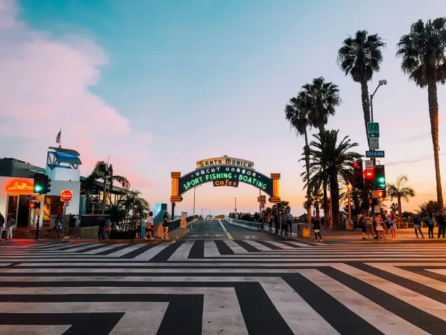 Santa Monica Pier entrance at sunset with palm trees and vibrant sky, bustling with visitors.